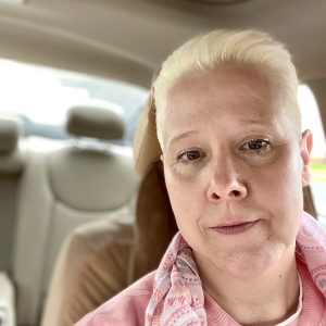 A white woman with blond hair is looking into the camera. The photo is taken inside a car. 