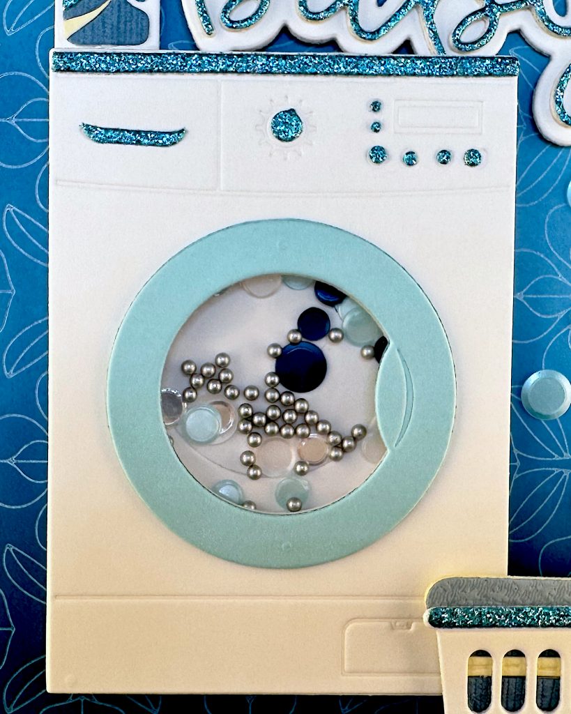 Close-up of the washing machine die cut with all the turquoise glitter details and the shaker window in the washing machine door. 