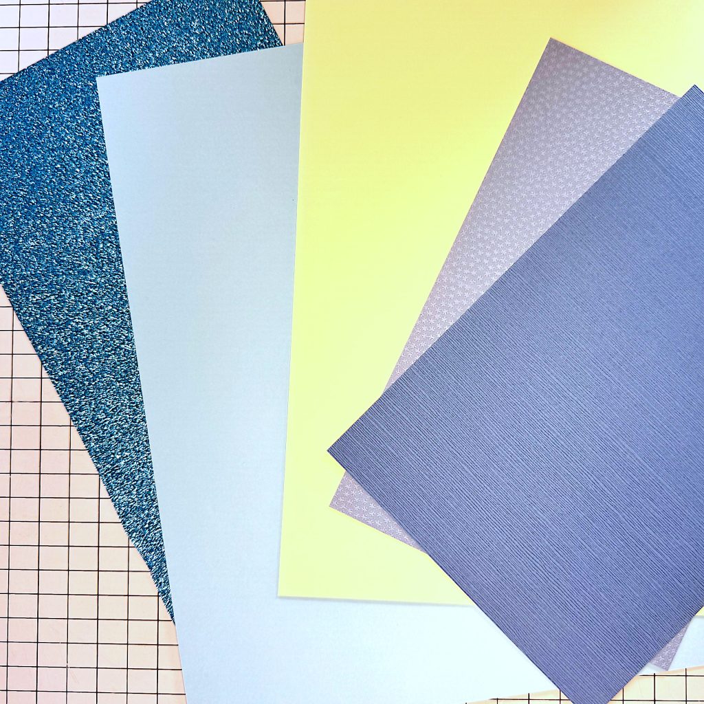 Five letter-sized pieces of cardstock. From left to right: Turquoise glitter cardstock, smooth sky blue cardstock, cream cardstock, light gray cardstock with a pebble texture and a darker blue gray cardstock with a linen texture. 