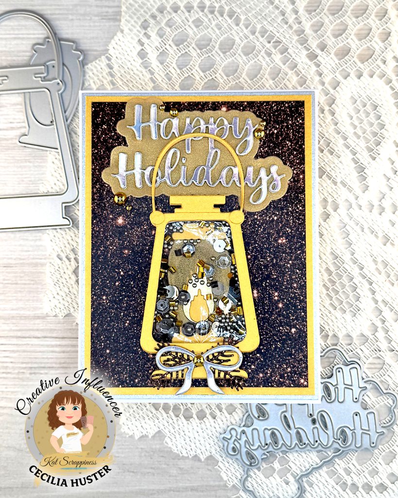 Greeting card in gold and silver tones. The main feature is a gold lantern with many gold and silver sequins inside. The large greeting reads "Happy Holidays." The card is lying on a piece of lace with some of the dies used to create it.
