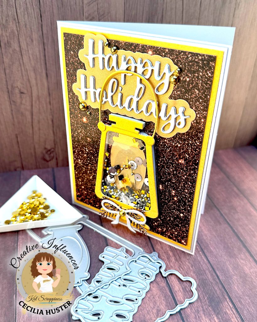 Greeting card in gold and silver tones. The main feature is a gold lantern with many gold and silver sequins inside. The large greeting reads "Happy Holidays." The card is standing up facing left. Some of the dies used to create it are lying in front of it, as is a triangle tray with gold flat back pearls.