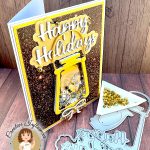 Greeting card in gold and silver tones. The main feature is a gold lantern with many gold and silver sequins inside. The large greeting reads "Happy Holidays." The card is standing up facing right. Some of the dies used to create it are lying in front of it, as is a triangle tray with gold flat back pearls.