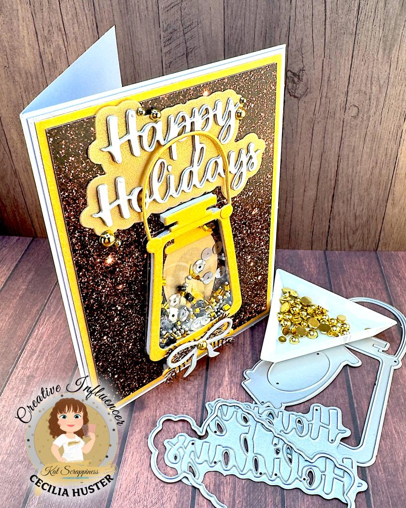 Greeting card in gold and silver tones. The main feature is a gold lantern with many gold and silver sequins inside. The large greeting reads "Happy Holidays." The card is standing up facing right. Some of the dies used to create it are lying in front of it, as is a triangle tray with gold flat back pearls.