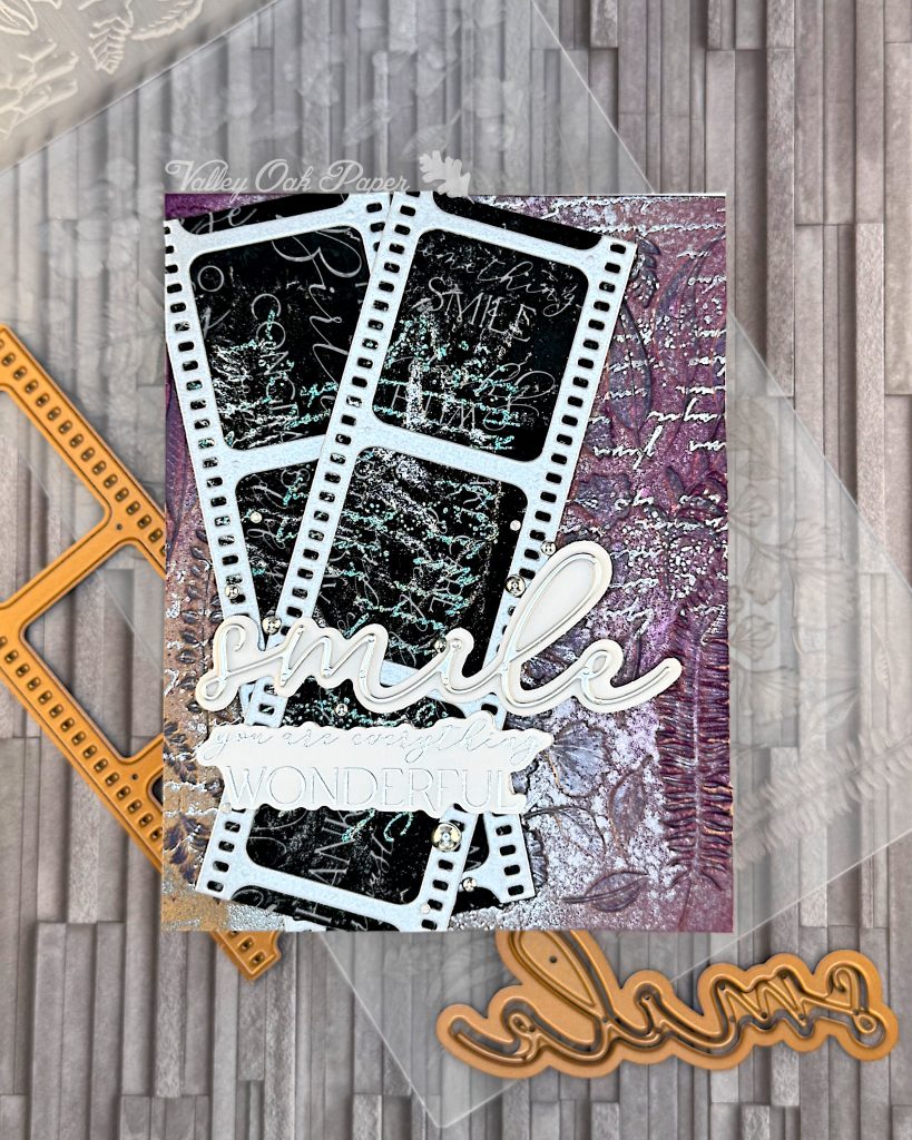 A mixed media greeting card is lying on an embossing folder and some copper-colored dies. The card has a purple and silver background with silver writing and botanical impressions. On top of the background there are two film strips with black, silver and green patterns and words in the images. On top of the film strips are two sentiments. One just says "smile," the other one says "You are everything wonderful."