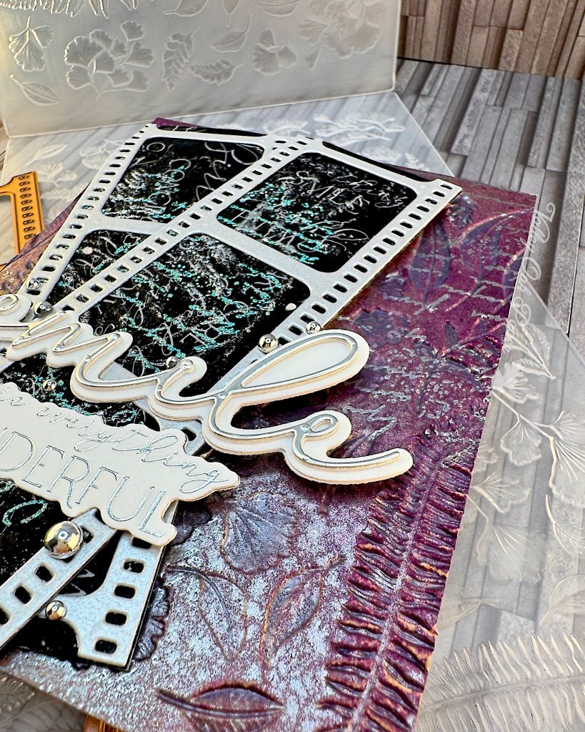 A closeup of a mixed media greeting card on an embossing folder. The card has a purple and silver background with silver writing and botanical impressions. On top of the background there are two film strips with black, silver and green patterns and words in the images. On top of the film strips are two sentiments. One just says "smile," the other one says "You are everything wonderful."