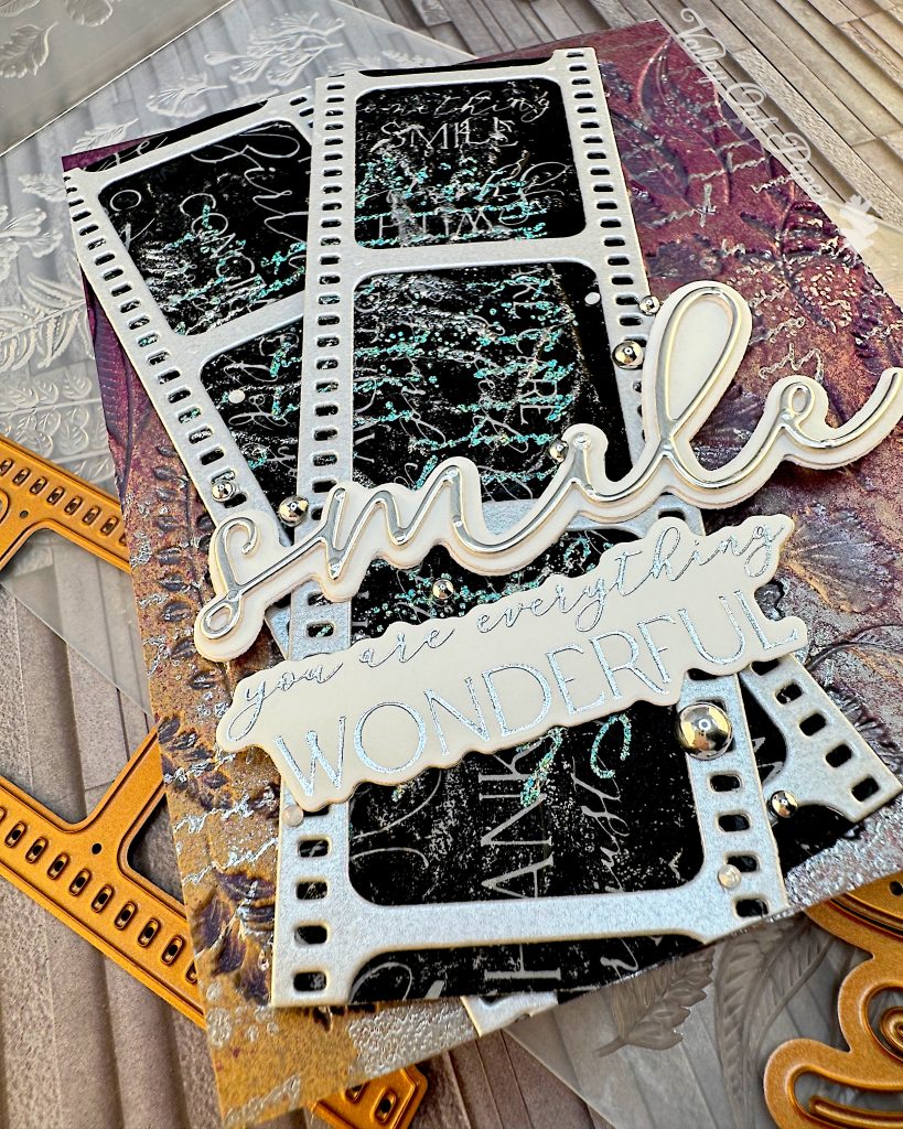 A closeup of a mixed media greeting card on an embossing folder. A few dies are also scattered around it. The card has a purple and silver background with silver writing and botanical impressions. On top of the background there are two film strips with black, silver and green patterns and words in the images. On top of the film strips are two sentiments. One just says "smile," the other one says "You are everything wonderful."