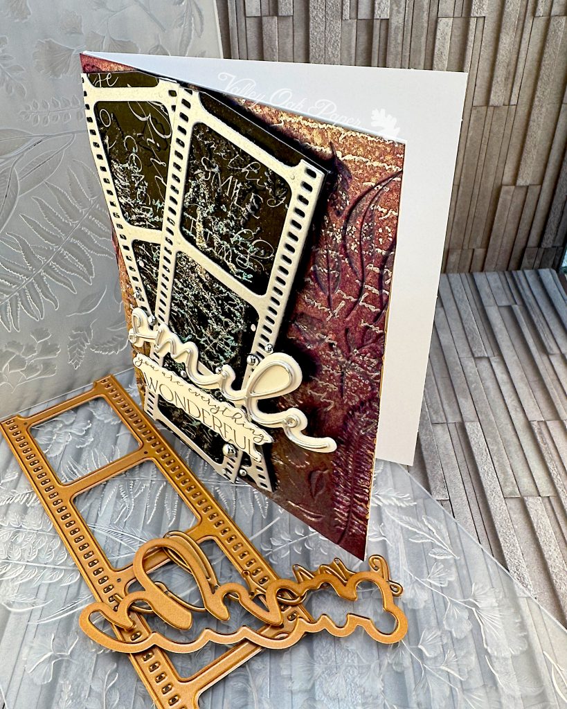 A mixed media greeting card stands on an embossing folder and some copper-colored dies. The card has a purple and silver background with silver writing and botanical impressions. On top of the background there are two film strips with black, silver and green patterns and words in the images. On top of the film strips are two sentiments. One just says "smile," the other one says "You are everything wonderful."