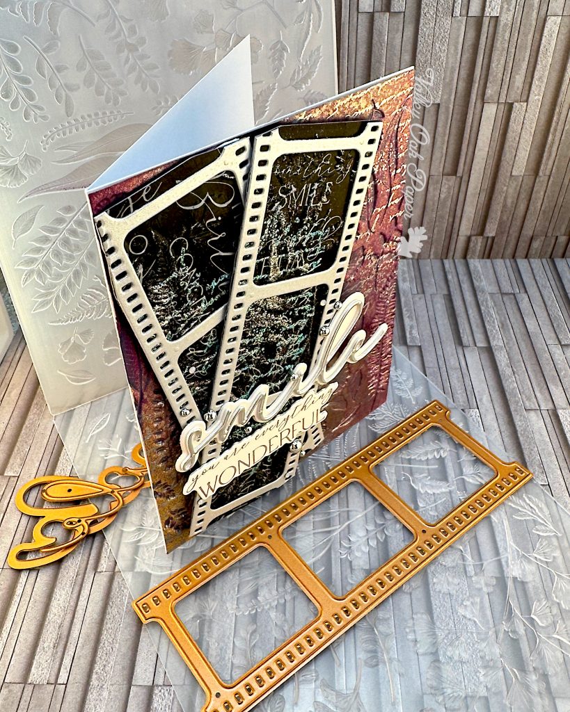 A mixed media greeting card stands on an embossing folder and some copper-colored dies. The card has a purple and silver background with silver writing and botanical impressions. On top of the background there are two film strips with black, silver and green patterns and words in the images. On top of the film strips are two sentiments. One just says "smile," the other one says "You are everything wonderful."