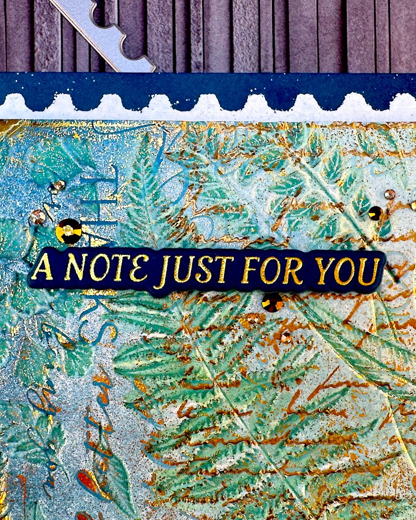 Closeup of the sentiment on a greeting card in blue and gold tones. The greeting reads "A note just for you." The background consists of several layers of color. You can make out the word THANKS. There are gold gems around the sentiment.