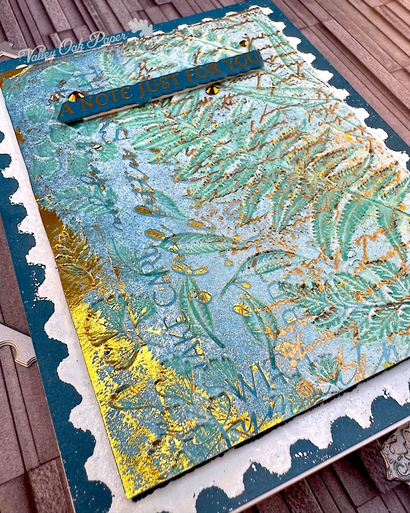 Greeting card in blue and gold tones on a slate grey background. The greeting reads "A note just for you." Closest to the camera is a part of the card that's mostly gold. You can make out the words "TAKE CARE."