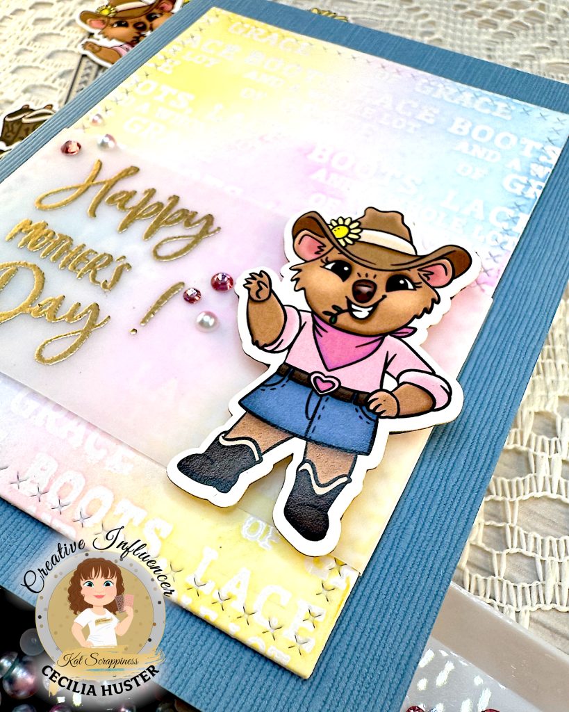Denim blue greeting card with a background in pastel colors. The focal point is a cowgirl quokka next to the sentiment. The sentiment reads "Happy Mother's Day!"
The card is lying at an angle, so that the cute cowgirl quokka is emphasized. She is wearing a denim skirt and a pink top.