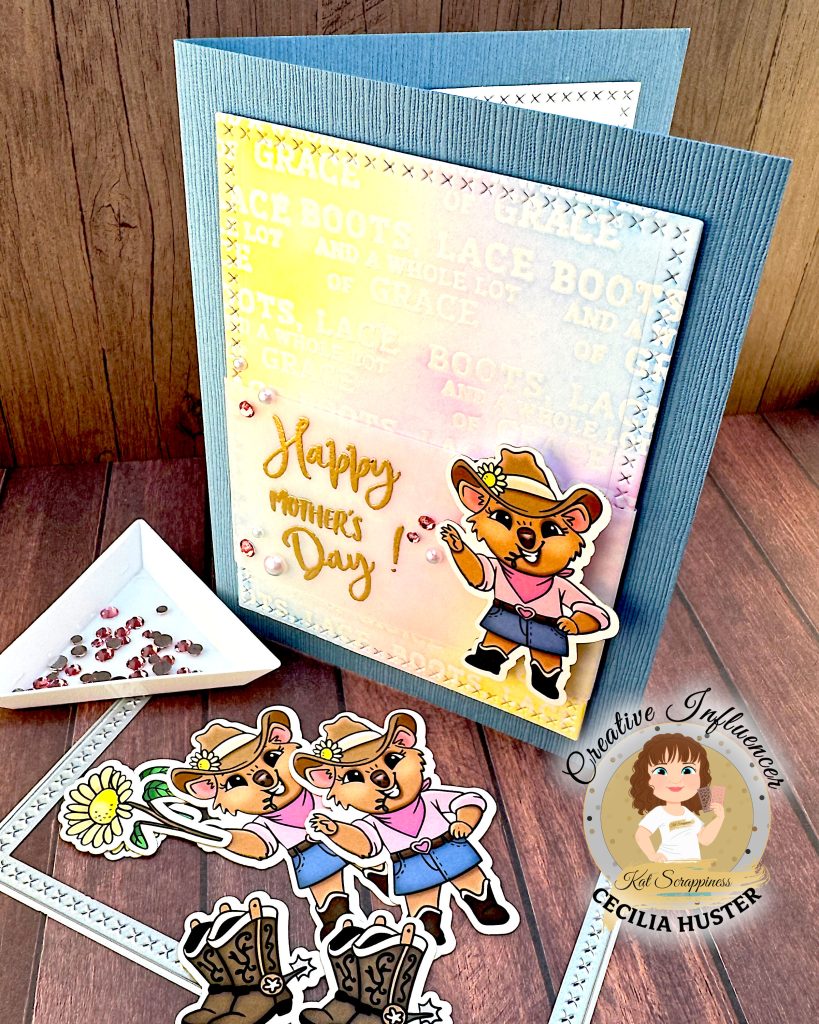 Denim blue greeting card is standing on a wooden surface. In front of it are more cowgirl quokka ephemera pieces, a cross stitch rectangle die and a triangle tray with pink sapphire rhinestones.