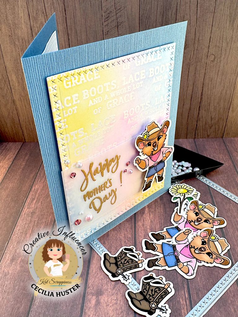 Denim blue greeting card is standing on a wooden surface. In front of it are more cowgirl quokka ephemera pieces, a cross stitch rectangle die and a black triangle tray with pink and blue pearls.