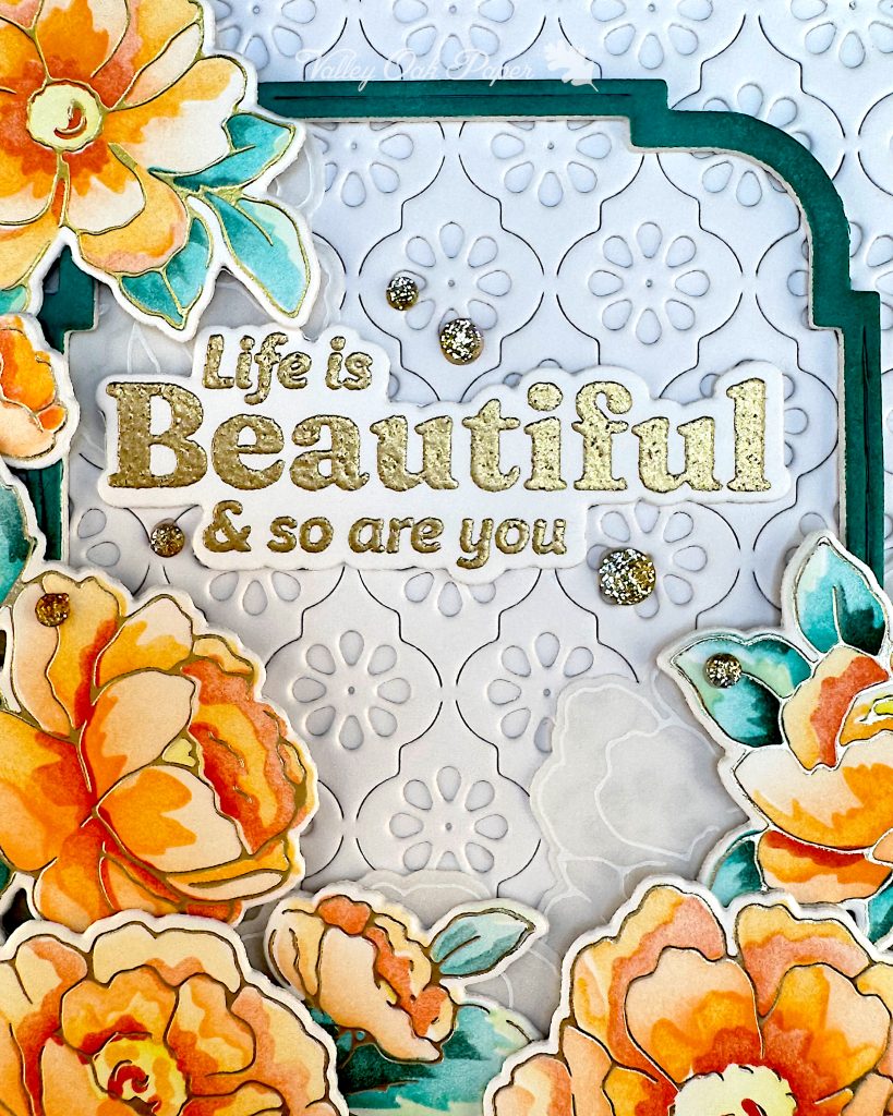 Detail from a white greeting card. The gold heat-embossed sentiment is the main focus. It reads "Life is beautiful and so are you." Gems are scattered around it. On the periphery you can see a green inset, rounded corner frame and flowers in sunset tones.