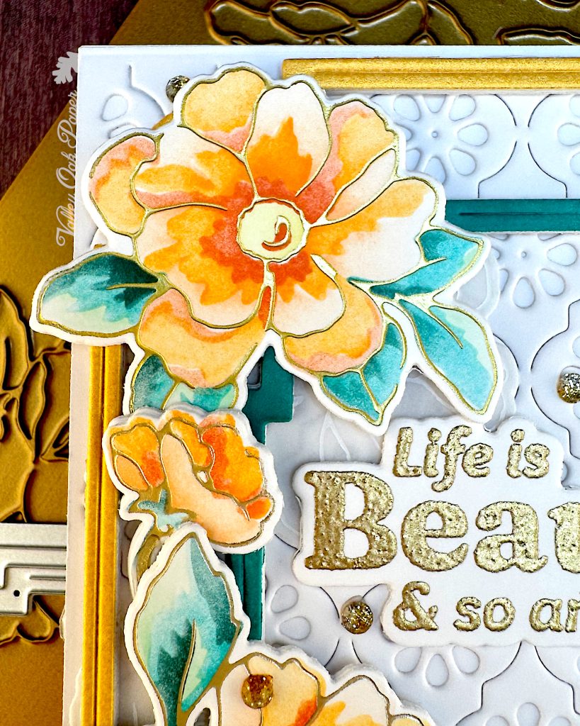 Detail from a white greeting card. The main focus is a large flower in the top right corner of the card. You can also make out the first few words in the gold sentiment.