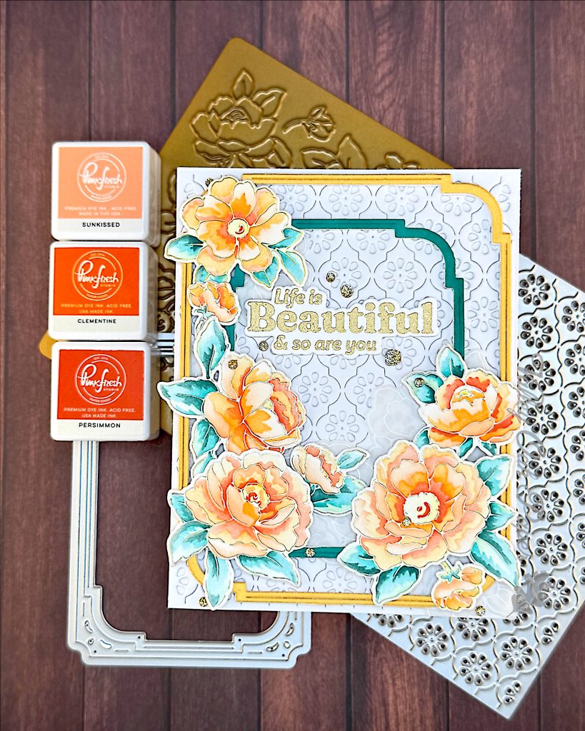 A white greeting card on a wooden background. There are two concentric frames, one gold, the other dark teal on the card. On top of those are peony flowers in sunset colors. The greeting reads "Life is beautiful and so are you."
In the background a floral press plate and a stylized flower pattern cover die are visible, as well as a frame die and three mini ink pads.