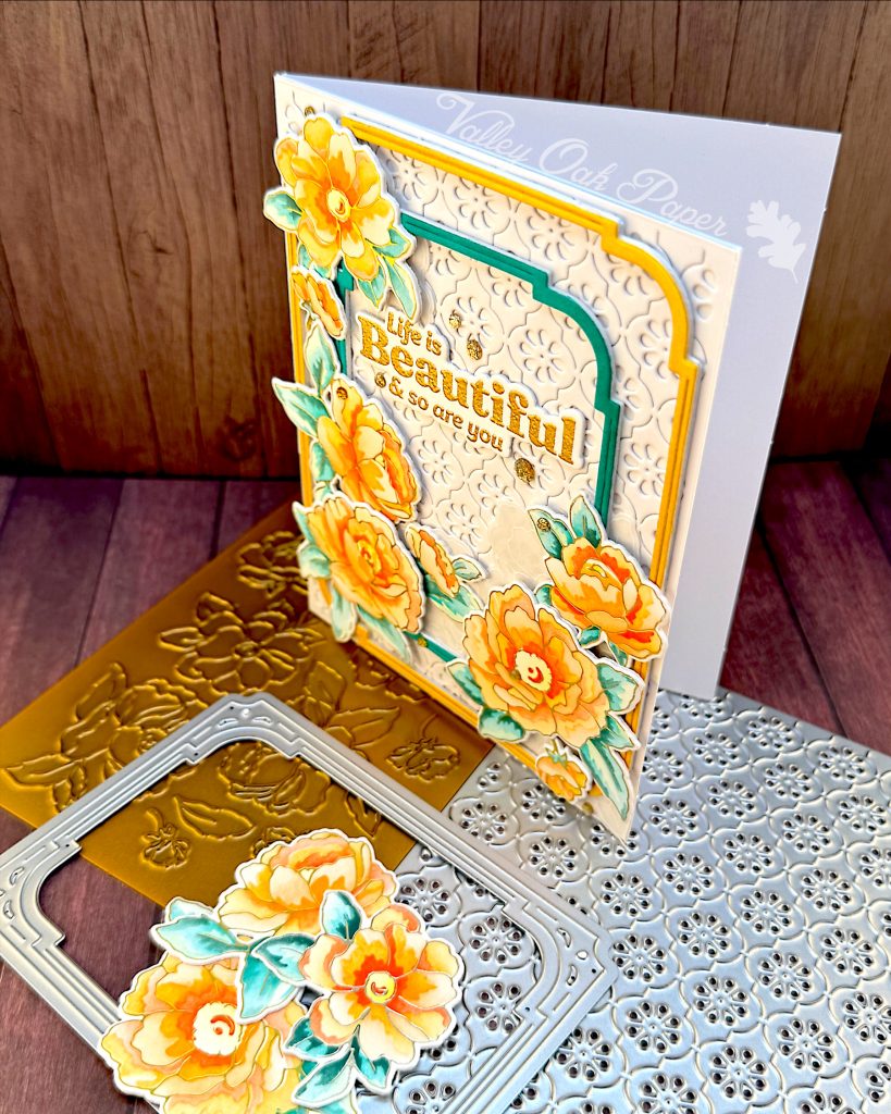 A white greeting card stands facing left on a wooden background. The card has a white textured background, on which lie two slim, rectangular frames with rounded corners. One in gold, the other dark teal. On top of those are peony flowers in sunset colors. The greeting reads "Life is beautiful and so are you."
The card stands on a floral press plate and a stylized flower pattern cover die.