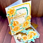 A white greeting card stands facing right on a wooden background. The card has a white textured background, on which lie two slim, rectangular frames with rounded corners. One in gold, the other dark teal. On top of those are peony flowers in sunset colors. The greeting reads "Life is beautiful and so are you." The card stands on a floral press plate and a frame die. In front of the card are some more flowers and sentiments in gold and dark teal.