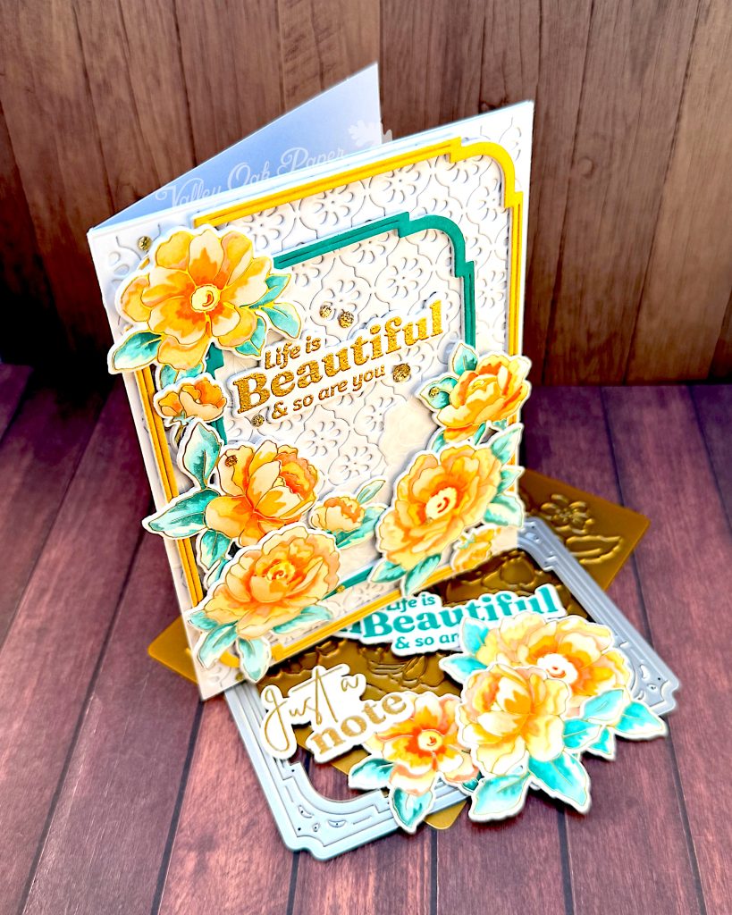 A white greeting card stands facing right on a wooden background. The card has a white textured background, on which lie two slim, rectangular frames with rounded corners. One in gold, the other dark teal. On top of those are peony flowers in sunset colors. The greeting reads "Life is beautiful and so are you."
The card stands on a floral press plate and a frame die. In front of the card are some more flowers and sentiments in gold and dark teal.