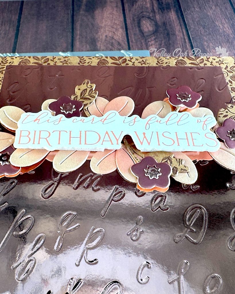 A greeting card in rosegold tones has been captured from an angle. From this vantage point you can tell that the main panel is rosegold mirror cardstock with an debossed pattern of individual cursive letters.
But it's the sentiment that stands out. It reads "This card is full of birthday wishes." It sits on top of some flowers and leaves cut out from cardstock in different rosegold tones and finishes.