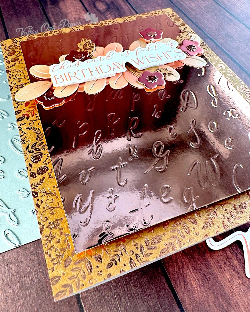 A greeting card in rosegold tones has been captured from an angle. The background is Kraft cardstock with a ditsy pattern in rosegold foil. The main panel is rosegold mirror cardstock with an embossed pattern of individual cursive letters. From this angle the beautiful mirror cardstock is highlighted.