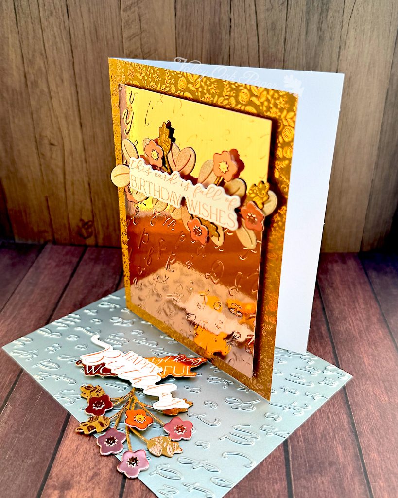A greeting card in rosegold tones. The background is Kraft cardstock with a ditsy pattern in rosegold foil. The main panel is rosegold mirror cardstock with an embossed pattern of cursive letters.
The greeting reads "This card is full of birthday wishes." It's anchored by leaves and flowers in different shades of rosegold.
The card is standing up and facing to the left. Under the card lies the pressplate that was used to create the letter pattern. There are also a few die cuts that didn't make it onto the card.