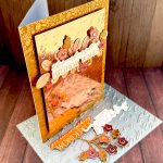 A greeting card in rosegold tones. The background is Kraft cardstock with a ditsy pattern in rosegold foil. The main panel is rosegold mirror cardstock with an embossed pattern of cursive letters. The greeting reads "This card is full of birthday wishes." It's anchored by leaves and flowers in different shades of rosegold. The card is standing up and facing to the right. Under the card lies the pressplate that was used to create the letter pattern. There are also a few die cuts that didn't make it onto the card.