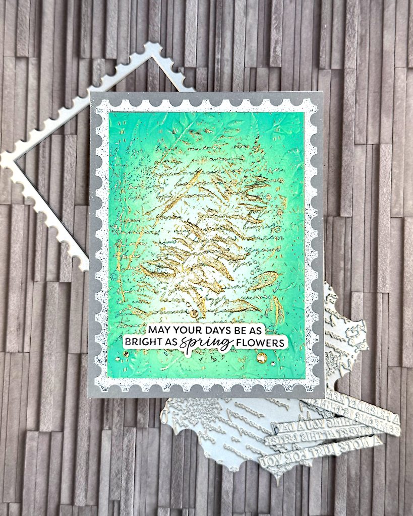 On a grey background lies a grey greeting card. A green and gold texture postage stamp is taking up most of the space. The greeting reads "May your days be as bright as spring flowers."
Under the card some of the press plates used to create it are peeking out.