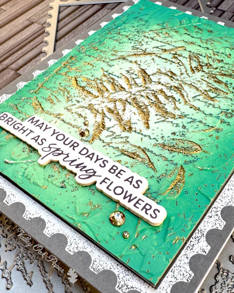 On a grey background lies a grey greeting card. It's angled so that the lower right corner makes up the foreground. You can clearly see the different textures and patterns.
The greeting reads "May your days be as bright as spring flowers." Under the card some of the press plates used to create it are peeking out.