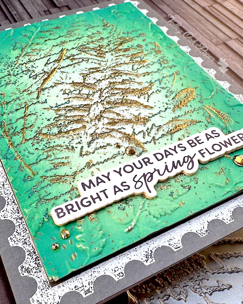 On a grey background lies a grey greeting card. It's angled so that the lower left corner makes up the foreground. You can clearly see the different textures and patterns.
The greeting reads "May your days be as bright as spring flowers." Under the card some of the press plates used to create it are peeking out.