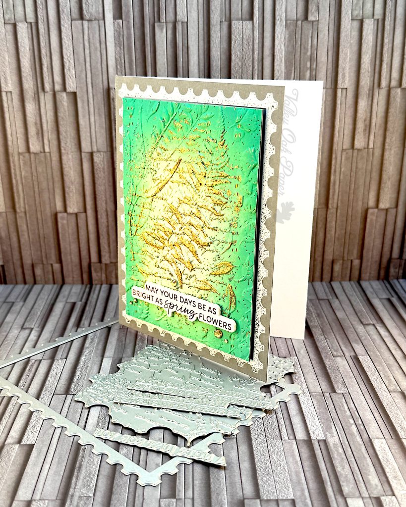 A grey greeting card facing left. A green and gold postage stamp is taking up most of the space. The greeting reads "May your days be as bright as spring flowers." The card is standing on some of the press plates used to create it.