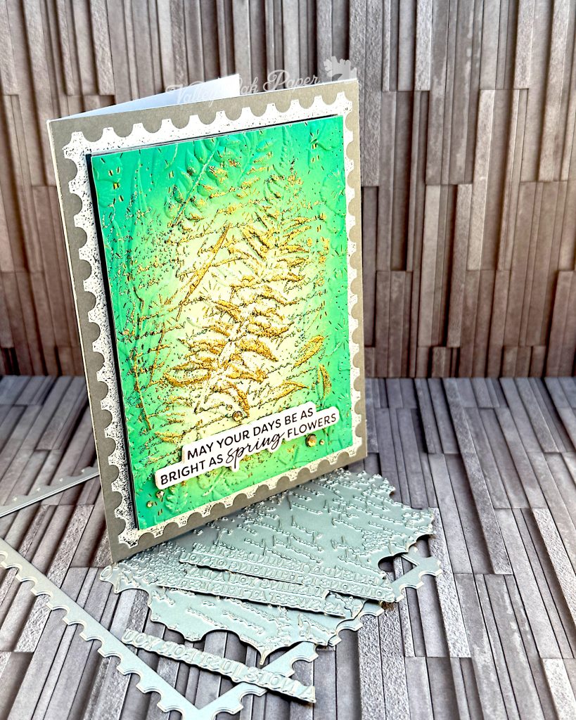 A grey greeting card facing right. A green postage stamp is taking up most of the space. The greeting reads "May your days be as bright as spring flowers." The card is standing on some of the press plates used to create it.
