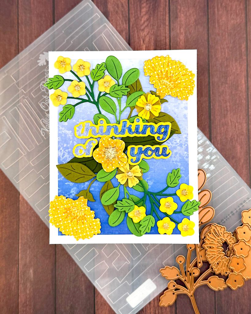 Greeting card with a blue gradient background and yellow flowers in a symmetrical pattern.
The greeting reads "Thinking of You" with a flower between the words.
Under the greeting card lies the embossing folder used in its creation and some floral dies.