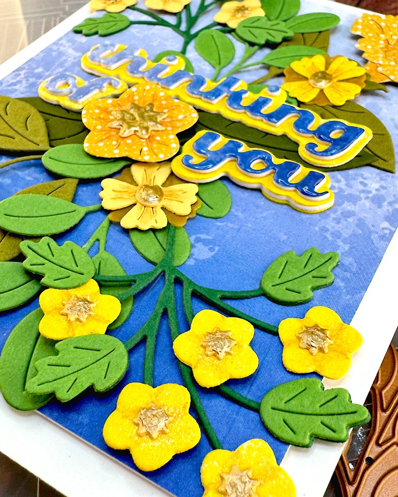 Angled shot of a greeting card with a blue gradient background and yellow flowers. The angle emphasizes the small yellow flowers with gold centers in the bottom right corner of the card.
The greeting reads "Thinking of You" with a flower between the words.