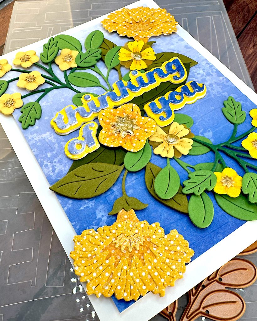 Angled shot of a greeting card with a blue gradient background and yellow flowers. The angle emphasizes the large trumpet-shaped yellow flower in the bottom left corner of the card.
The greeting reads "Thinking of You" with a flower between the words.