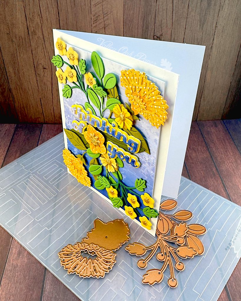 A greeting card with a blue gradient background and yellow flowers in a symmetrical pattern.
The greeting reads "Thinking of You" with a flower between the words.
The card is standing, facing left, on an embossing folder used in its creation. A few floral dies are scattered about.