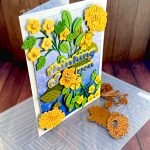A greeting card with a blue gradient background and yellow flowers in a symmetrical pattern. The greeting reads "Thinking of You" with a flower between the words. The card is standing, facing right, on an embossing folder used in its creation. A few floral dies are scattered about.