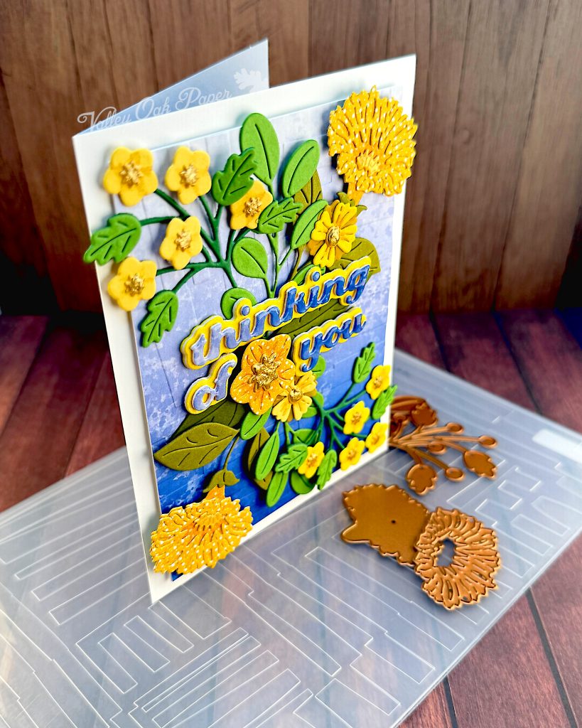 A greeting card with a blue gradient background and yellow flowers in a symmetrical pattern.
The greeting reads "Thinking of You" with a flower between the words.
The card is standing, facing right, on an embossing folder used in its creation. A few floral dies are scattered about.