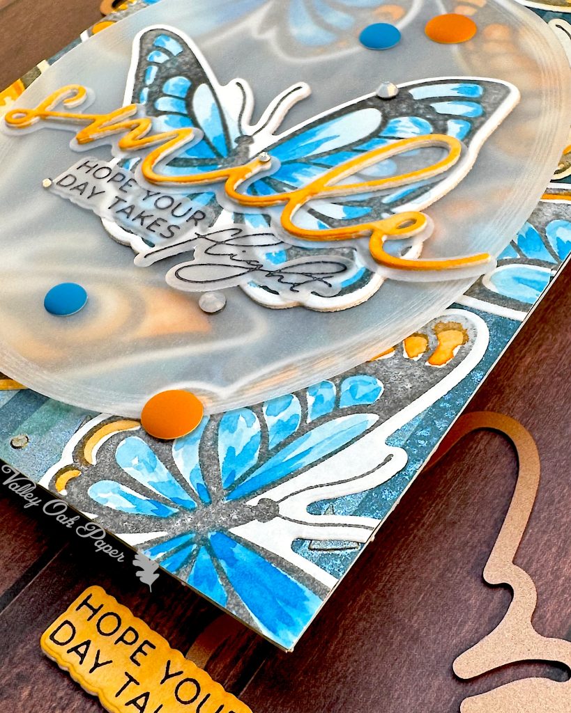 Angled view of a card featuring big butterflies in orange and blue tones. The focal point is a blue butterfly. The main greeting reads "smile." It sits across the blue butterfly. Under it the subsentiment says "Hope your day takes flight."
The angle highlights some of the orange and blue enamel dots used to decorate the card.