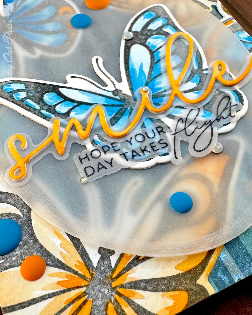 Close up of a card featuring big butterflies in orange and blue tones. The focal point is a blue butterfly. The main greeting reads "smile." It sits across the blue butterfly. Under it the subsentiment says "Hope your day takes flight."
The close up emphasizes the blue and orange matte enamel dots used to embellish the card.