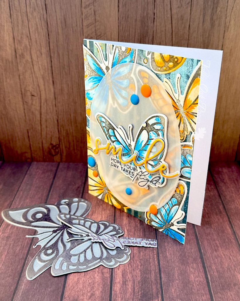 Greeting card featuring big butterflies in orange and blue tones. The focal point is a blue butterfly. The main greeting reads "smile." It sits across the blue butterfly. Under it the subsentiment says "Hope your day takes flight."
The card stands on a wood background with some of the BetterPress plates used to create it in front of it.