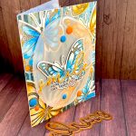 Greeting card featuring big butterflies in orange and blue tones. The focal point is a blue butterfly. The main greeting reads "smile." It sits across the blue butterfly. Under it the subsentiment says "Hope your day takes flight." The card stands on a wood background with the "smile" die set used to create the sentiment in front of it.