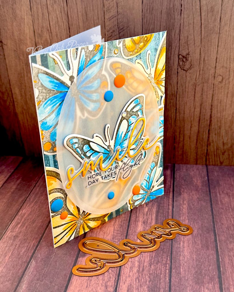 Greeting card featuring big butterflies in orange and blue tones. The focal point is a blue butterfly. The main greeting reads "smile." It sits across the blue butterfly. Under it the subsentiment says "Hope your day takes flight."
The card stands on a wood background with the "smile" die set used to create the sentiment in front of it.