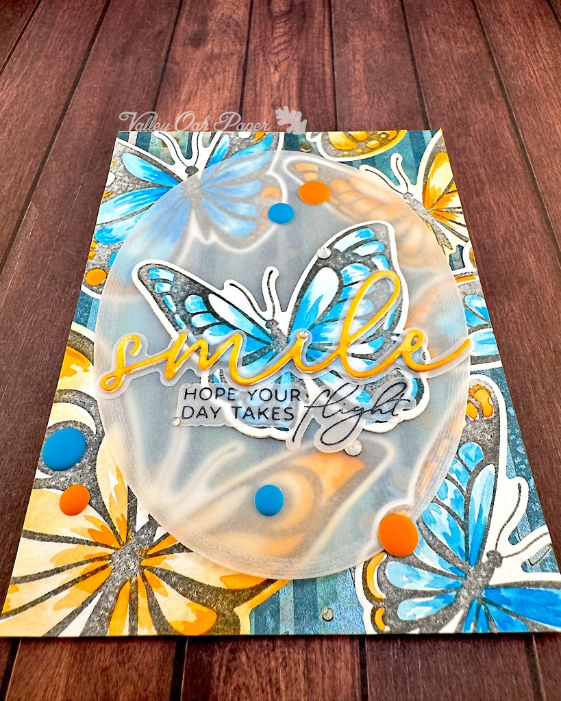 Greeting card featuring big butterflies in orange and blue tones. The focal point is a blue butterfly. The main greeting reads "smile." It sits across the blue butterfly. Under it the subsentiment says "Hope your day takes flight."
The card is lying on a wood background in warm tones.