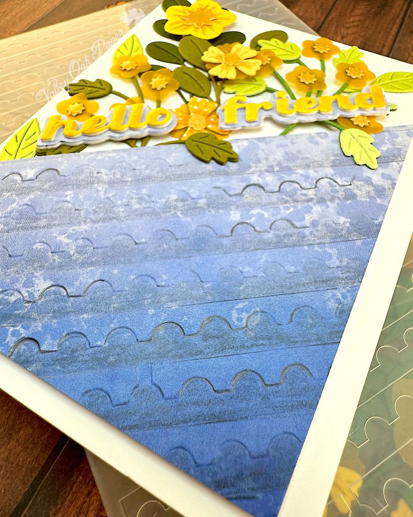 An angled shot of a white greeting card with half of the surface a blue gradient embossed with a postage stamp edge design. The angle makes the pattern stand out. The flowers and foliage disappear into the background.