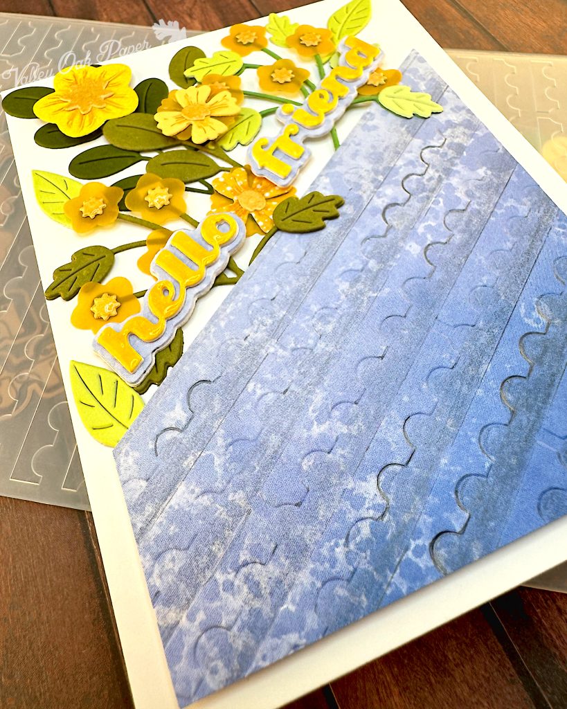 An angled shot of a white greeting card with half of the surface a blue gradient embossed with a postage stamp edge design. The angle makes the pattern stand out. The flowers and foliage disappear into the background.