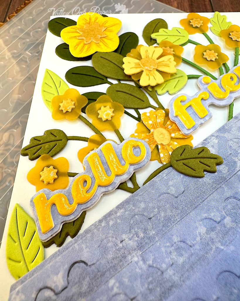 Closeup of the word "hello" on a greeting card. The word is yellow and shiny. The font is a chunky script font. Under the word is a light blue shadow layer. Under that are flowers in yellow tones.
The word has been placed at an angle, so that it's long edge is parallel with the edge of a blue piece of cardstock.