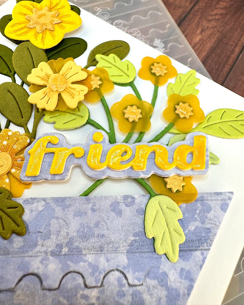 Closeup of the word "friend" on a greeting card. The word is yellow and shiny. The font is a chunky script font. Under the word is a light blue shadow layer. Under that are flowers in yellow tones.
The word has been placed at an angle, so that it's long edge is parallel with the edge of a blue piece of cardstock.