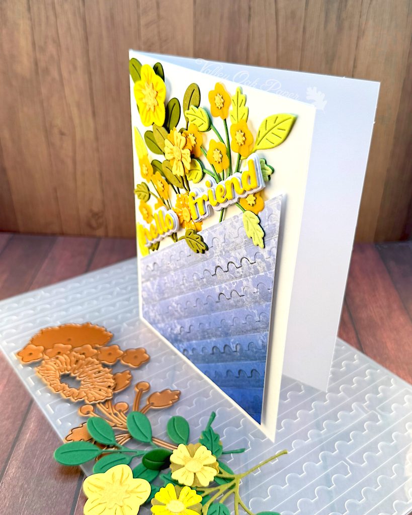White greeting card with half of the surface a blue gradient embossed with a postage stamp edge design. The other half is a mix of flowers and foliage in yellow and green tones. The sentiment reads "Hello Friend."
The card is standing up, facing left, on the postage edge embossing folder that was used to create the blue panel. Some more yellow flowers and dies are lying in front of it.