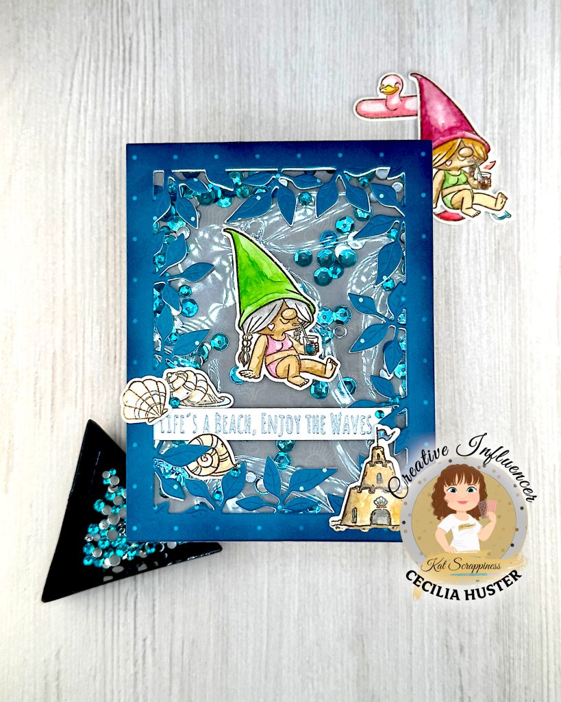 A handmade greeting card with a beach gnome theme displayed against a pale wooden surface. The card features a clear window filled with turquoise and silver sequins. The window is bordered by a teal, leafy frame.
The main design shows a cute gnome with long, grey hair and a bright green pointed hat. She appears to be relaxing in a bikini while holding a small drink. A banner across the card reads "LIFE'S A BEACH, ENJOY THE WAVES!" The banner is decorated with sea shells. A sandcastle has been placed in the lower right corner.
Under the card are several coordinating die-cut elements including another gnome figure with a pink hat and a pink flamingo pool floatie. A black triangle tray holding turquoise gems is also visible.
The bottom right corner shows the "Kat Scrappiness Creative Influencer CECILIA HUSTER" logo featuring a cartoon woman with brown hair.